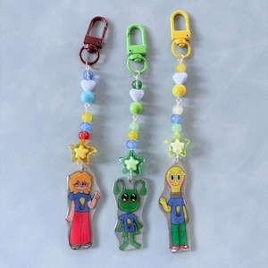Puede incluir: Tres llaveros de acrílico con personajes de dibujos animados que llevan iconos de bombillas en sus camisetas. Cada llavero tiene una cadena de cuentas coloridas con formas de corazón y estrella, unida a un mosquetón de metal.
