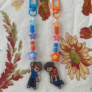 May include: Two keychains featuring cartoon character charms. One has a blue lobster clasp with blue and orange star and round beads. The other has an orange lobster clasp with orange, pink, and blue star and round beads. Each charm depicts a person.