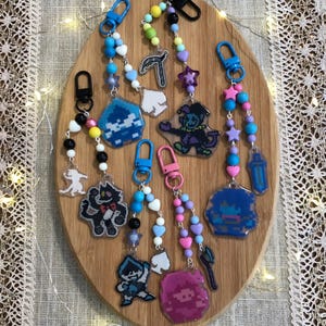 May include: A collection of colorful beaded keychains with various character charms. The keychains feature blue, pink, and purple beads, with charms depicting pixelated characters and shapes. Each keychain has a metal clasp.