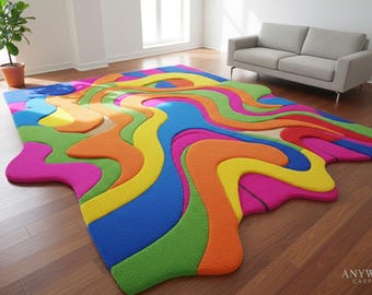 May include: A vibrant, abstract area rug with a colorful, flowing design. The rug features a mix of bright colors including pink, yellow, green, blue, and orange. The rug is placed on a hardwood floor, with a gray sofa in the background.