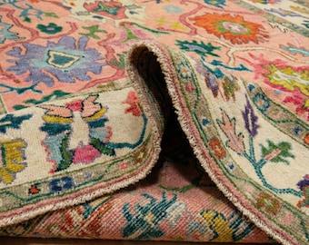 Könnte beinhalten: Aufgerollter Teppich mit detailliertem Blumenmuster. Der Teppich hat einen hellrosa Hintergrund mit farbenfrohen Blumenmotiven in Blau, Orange, Grün und Lila. Der Rand ist cremefarben mit grüner Umrandung.