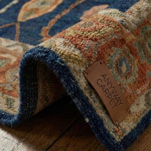 Peut inclure: Gros plan sur un tapis roulé avec un motif floral détaillé dans les tons de bleu, orange et beige. Le tapis a une bordure bleu foncé et une étiquette en cuir qui dit "ANYWAY CARPET".