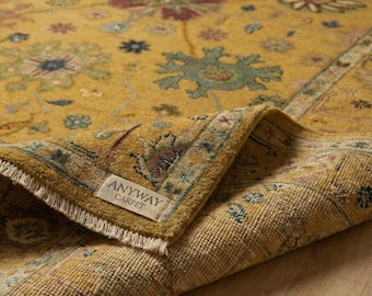 May include: A mustard yellow area rug featuring a repeating floral pattern in shades of blue, green, and maroon. The rug has a folded corner revealing a white fabric label with the text ANYWAY CARPET and a short white fringe edge.