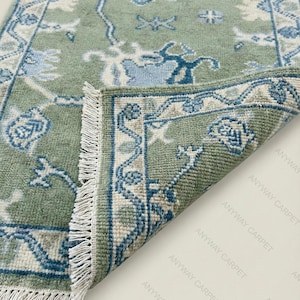 May include: A sage green rug with a detailed floral pattern in shades of blue and cream. The rug has a fringed edge and a folded corner, showcasing the intricate design. The rug is a decorative home accent.