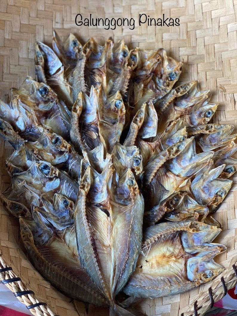 May include: A woven basket filled with dried, golden brown fish. The fish are arranged in a circular pattern, with their heads pointing outwards. The text "Galunggong Pinakas" is written in black letters at the top of the image.