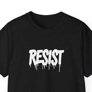 Puede incluir: Camiseta negra con un gráfico blanco "RESIST" en un estilo de pintura que gotea.