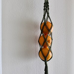 May include: A green macrame hanging fruit basket with three oranges inside. The basket is hanging from a brass hook on a white wall.