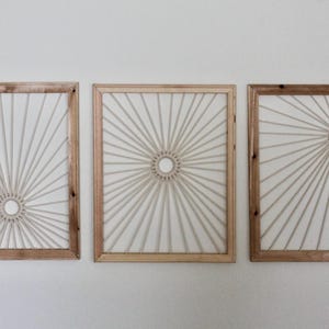 May include: Three framed wall art pieces. Each features a sunburst design made of off-white rope against a white background. The frames are made of light brown wood, creating a natural, neutral aesthetic. The art is mounted on a white wall.