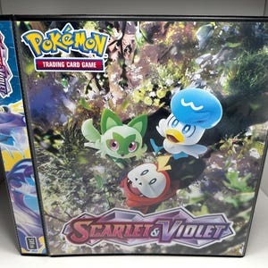 May include: A black binder with a Pokemon Scarlet and Violet theme. The binder features a colorful illustration of three Pokemon characters, including a green and white Sprigatito, a blue and white Quaxly, and a red and white Fuecoco. The binder has the text "Pokemon Trading Card Game" and "Scarlet & Violet" on the front.