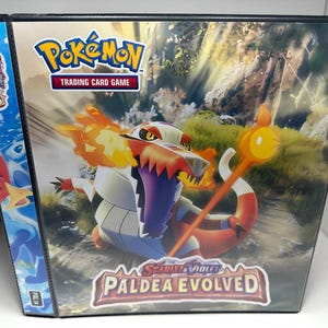 May include: A black binder with a colorful Pokemon Trading Card Game image on the front. The image features a fiery orange and red Pokemon with a white and purple body. The text "Scarlet & Violet Paldea Evolved" is printed on the front of the binder.