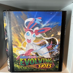 May include: A black binder with a colorful image of a pink and blue Pokemon character on the front. The text "SWORD & SHIELD EVOLVING SKIES" is printed on the front of the binder.