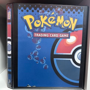 May include: A blue Pokemon Trading Card Game binder with a black border and a Pokeball design. The Pokeball is red, white, and black with a blue ring around the center.