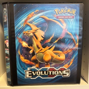 May include: A black binder with a colorful Pokemon card image on the front. The image features a red and orange dragon-like Pokemon with flames in the background. The text "Pokemon Trading Card Game" and "Evolutions" are visible on the binder.