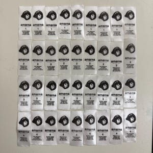 Puede incluir: Un conjunto de etiquetas de tela blanca con texto negro y una ilustración negra de una persona con sombrero. Las etiquetas están dispuestas en filas y columnas. El texto incluye la marca "amarna studios" y los números 1-4 con citas.