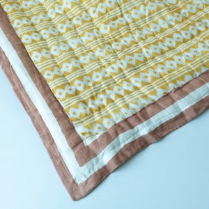 May include: A yellow and white patterned quilt with a brown and white border. The quilt is folded in half and the corner is visible.