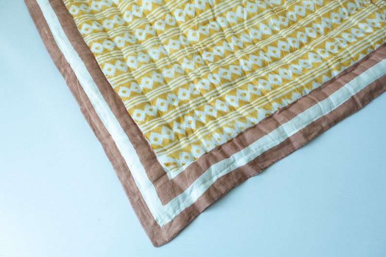 May include: A yellow and white patterned quilt with a brown and white border. The quilt is folded in half and the corner is visible.
