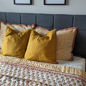 May include: A bed with a gray headboard and a white and brown floral patterned quilt. There are three pillows on the bed, two are mustard yellow and one has a white and yellow geometric pattern. Two framed pictures are hanging on the wall above the headboard.