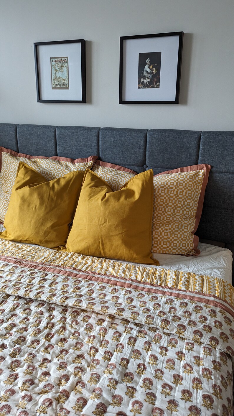 May include: A bed with a gray headboard and a white and brown floral patterned quilt. There are three pillows on the bed, two are mustard yellow and one has a white and yellow geometric pattern. Two framed pictures are hanging on the wall above the headboard.