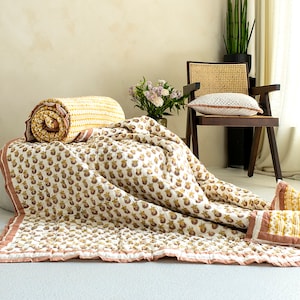 May include: A white and yellow floral patterned quilt with a pink border. The quilt is folded and laid out on a light brown surface. A small bouquet of pink flowers is in the background.