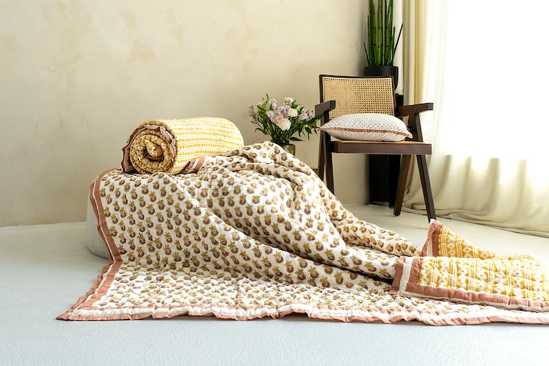 May include: A white and yellow floral patterned quilt with a pink border. The quilt is folded and laid out on a light brown surface. A small bouquet of pink flowers is in the background.