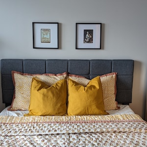 May include: A bedroom with a gray upholstered headboard, two yellow throw pillows, and a patterned quilt. There are two wooden dressers with drawers on either side of the bed. The walls are painted a light gray and there are two framed pictures above the headboard.