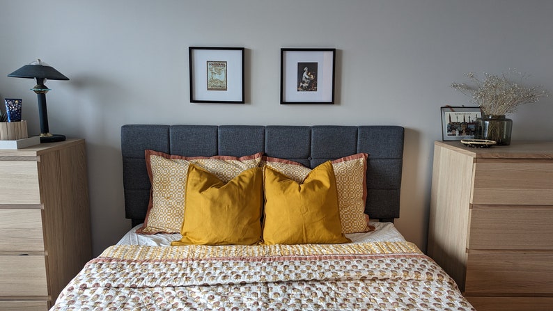 May include: A bedroom with a gray upholstered headboard, two yellow throw pillows, and a patterned quilt. There are two wooden dressers with drawers on either side of the bed. The walls are painted a light gray and there are two framed pictures above the headboard.
