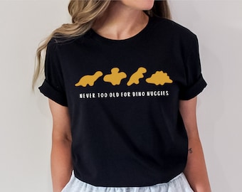 May include: Black t-shirt with a white graphic of four dinosaur-shaped chicken nuggets and the text "NEVER TOO OLD FOR DINO NUGGIES".
