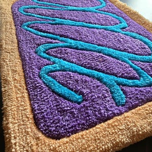 May include: A rectangular rug with a purple center, light blue squiggly lines, and a tan border. The rug has a plush, textured surface, resembling a frosted pastry. The design is reminiscent of a breakfast treat.