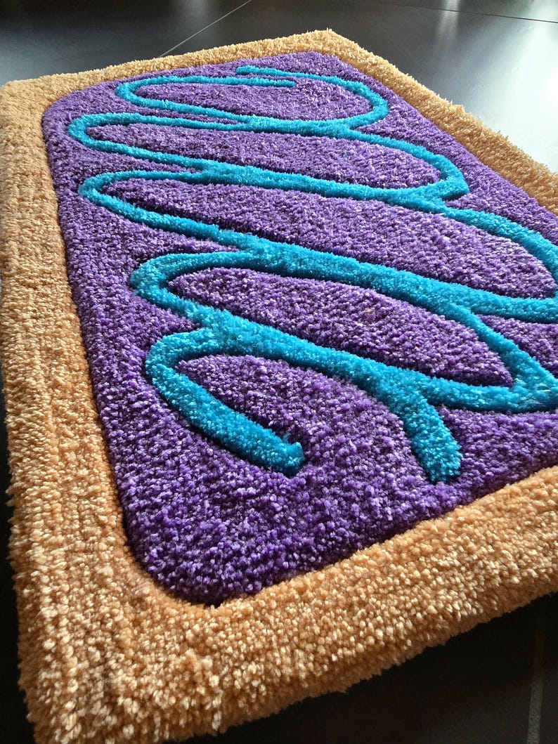 May include: A rectangular rug with a purple center, light blue squiggly lines, and a tan border. The rug has a plush, textured surface, resembling a frosted pastry. The design is reminiscent of a breakfast treat.