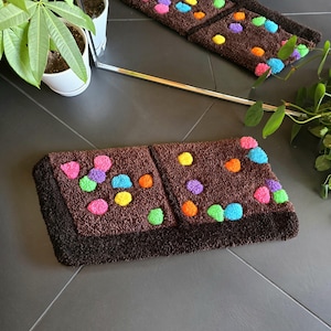 May include: A brown bath mat with a chocolate bar design. The mat has a dark brown border and is decorated with colorful sprinkles.