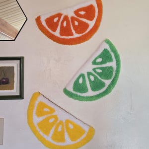 May include: Three fruit slice rugs in orange, green, and yellow. The rugs depict an orange, a lime, and a lemon, each with a white rind and a tufted design. They are mounted on a white wall.