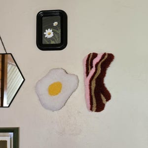 May include: A wall-mounted art arrangement featuring a small black framed picture with daisies, a fried egg rug with a yellow yolk, and a bacon rug with pink and brown stripes. The items are displayed on a neutral-colored wall.