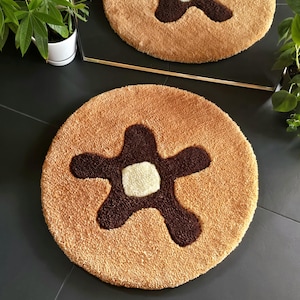 Può includere: Due tappetini da bagno rotondi e morbidi. Ogni tappetino ha uno sfondo beige con un disegno a forma di stella marrone scuro al centro e un piccolo quadrato bianco al centro della stella. I tappetini sono su una superficie grigio scuro.