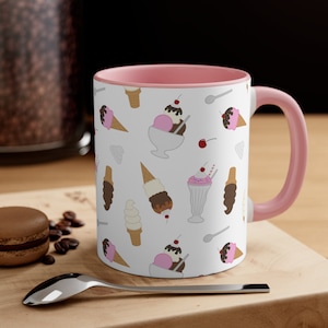 May include: A pink and white ceramic mug with a pattern of ice cream cones, sundaes, and milkshakes. The mug has a pink interior and a white exterior.