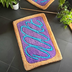 May include: A rectangular bath mat with a purple center, teal squiggles, and a tan border. The mat is designed to resemble a frosted pastry. The mat is made of a soft, plush material.