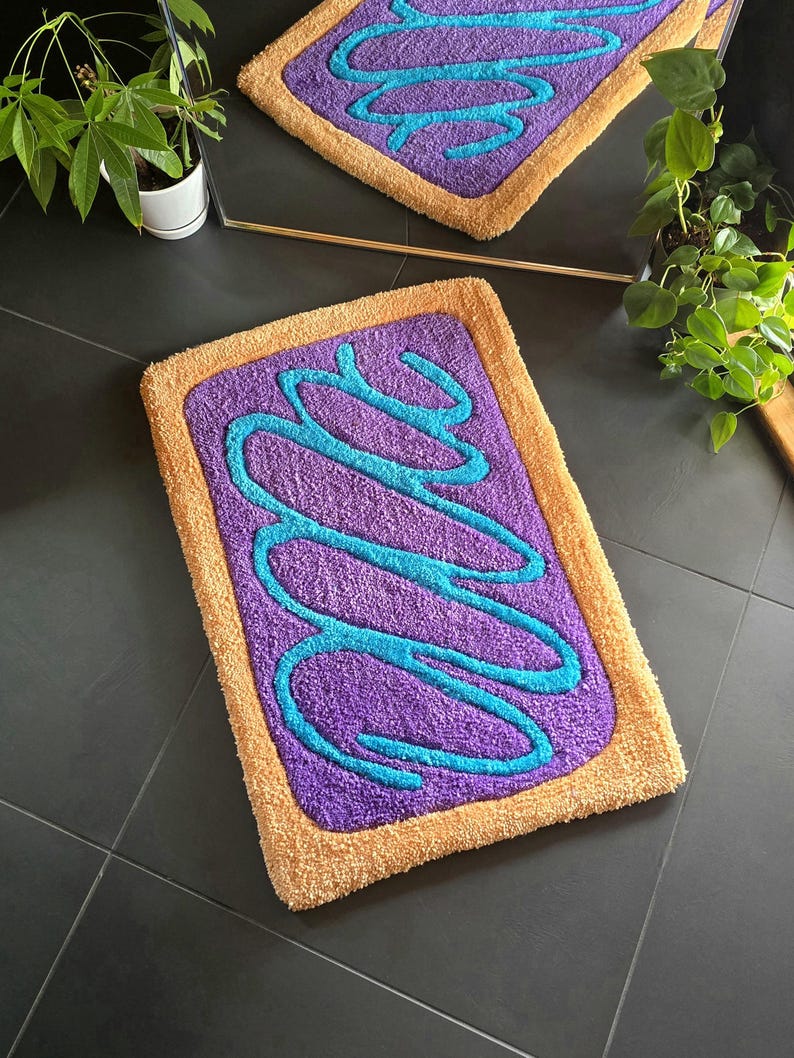May include: A rectangular bath mat with a purple center, teal squiggles, and a tan border. The mat is designed to resemble a frosted pastry. The mat is made of a soft, plush material.