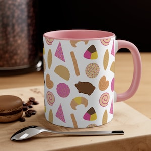 May include: A pink and white ceramic mug featuring a pattern of assorted Mexican pastries and sweets. The mug has a pink interior and a white exterior.