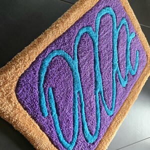 May include: A rectangular rug with a purple center and a tan border. The center features a teal-colored, cursive word design. The rug has a plush, textured appearance, suggesting a soft, comfortable feel. The design is reminiscent of a popular breakfast pastry.
