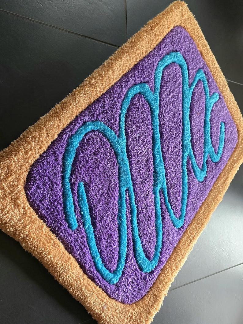 May include: A rectangular rug with a purple center and a tan border. The center features a teal-colored, cursive word design. The rug has a plush, textured appearance, suggesting a soft, comfortable feel. The design is reminiscent of a popular breakfast pastry.