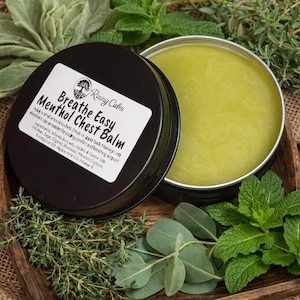 May include: A black tin of "Breathe Easy Menthol Chest Balm" with the lid off, revealing a light green balm. The tin sits in a wooden tray surrounded by fresh herbs. The label includes the brand name "Rising Calm".