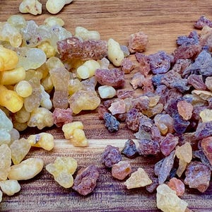 May include: Close-up of two piles of natural resin. One pile is a mix of yellow, amber, and clear pieces. The other pile is a mix of purple, pink, and brown pieces. The resin pieces are on a wooden surface.