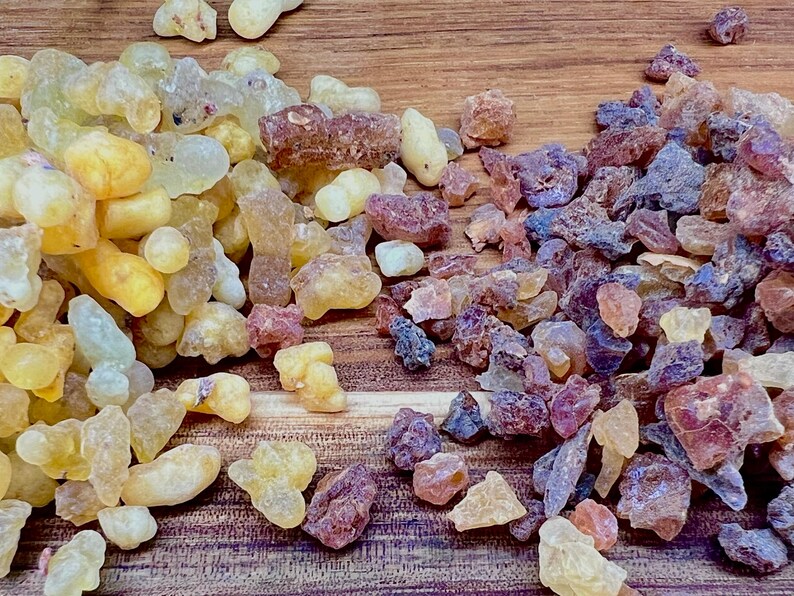 May include: Close-up of two piles of natural resin. One pile is a mix of yellow, amber, and clear pieces. The other pile is a mix of purple, pink, and brown pieces. The resin pieces are on a wooden surface.