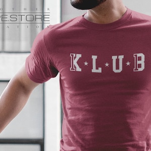 Op de afbeelding: Een maroon t-shirt met de tekst "K*L*U*B" in witte letters.