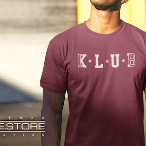 Op de afbeelding: Een maroon t-shirt met een witte afbeelding die "K*L*U*B" zegt.