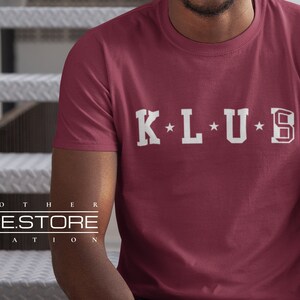 Op de afbeelding: Een maroon t-shirt met een witte afbeelding met de tekst "K*L*U*B" in een vintage universitaire stijl.