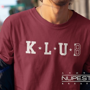 Op de afbeelding: Bordeauxkleurig T-shirt met ronde hals en het woord "KLUB" in witte blokletters. De letters "K", "L" en "U" hebben elk een ster. De letter "B" staat in een kader. De tekst "ANOTHER NUPE.STORE KREATION" staat onderaan.