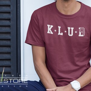 Op de afbeelding: Een maroon t-shirt met witte tekst die "K*L*U*B" zegt.