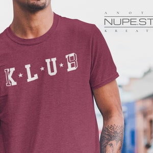Op de afbeelding: Een maroon t-shirt met witte tekst die "K*L*U*B" luidt. De tekst is in een vintage universitaire stijl lettertype. Het shirt is een ronde hals stijl en heeft korte mouwen.