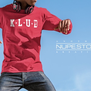 Op de afbeelding: Een rood longsleeve shirt met witte tekst die "K*L*U*B" op de voorkant staat. Het shirt wordt gedragen door een persoon met een koptelefoon op. De tekst "ANOTHER NUPEST.STORE KREATION" is zichtbaar in de rechterbenedenhoek van de afbeelding.