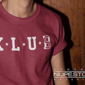 Op de afbeelding: Een maroon t-shirt met witte letters die "K*L*U*B" lezen. Het shirt is een crew neck-stijl en heeft opgerolde mouwen. De tekst "ANOTHER NUPEST.STORE KREATION" is zichtbaar in de rechterbenedenhoek van de afbeelding.